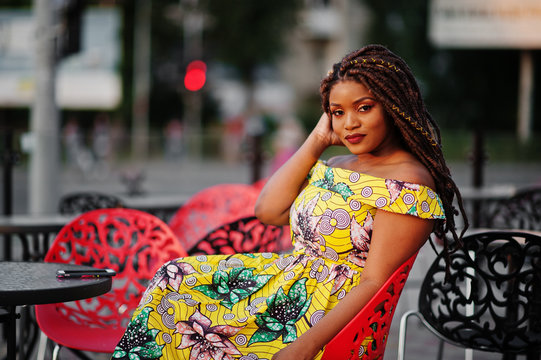 Cute small height african american girl with dreadlocks, wear at coloured yellow dress, sitting at outdoor cafe on red chair.