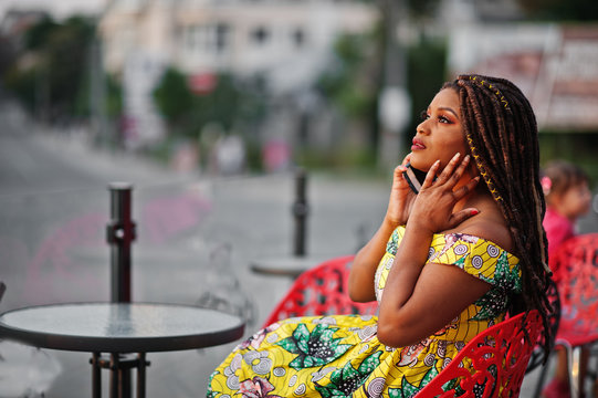 Cute small height african american girl with dreadlocks, wear at coloured yellow dress, sitting at outdoor cafe on red chair and speaking on phone.