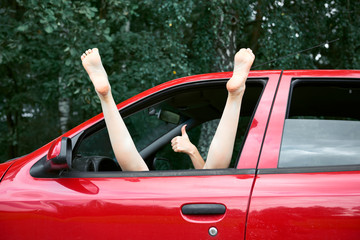 young woman driver resting in a red car, put her feet on the car window and gesturing, happy travel...