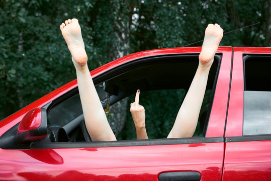 young woman driver resting in a red car, put her feet on the car window and shows the indecent gesture with middle finger
