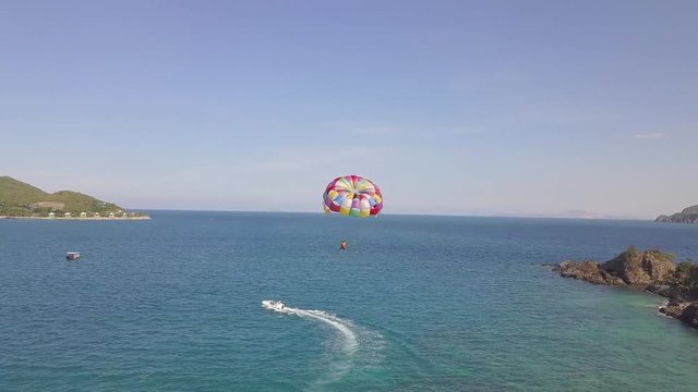 Aerial view parasail flying over blue sea pulled boat. Parasailing in sea bay and green island landscape drone view. Summer activity and extreme entertainment while resting on resort beach.