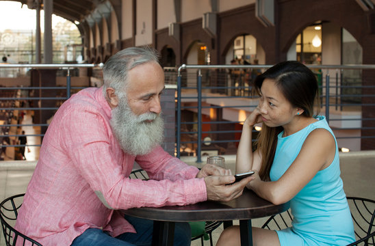 Lovely couple using smart phone looking at mobile smiling. Bearded senior man and young asian woman.