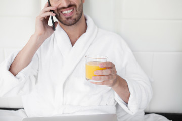 Young man having breakfast and talking on phone in bed. Indoors.