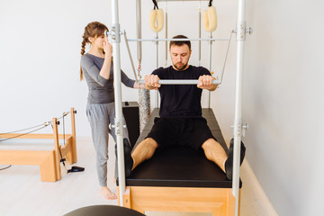 Young sporty female instructor of pilates helping beard man workout in cadillac bed. Two people working in pilates studio, woman assistant supporting and correcting male patient beginner.