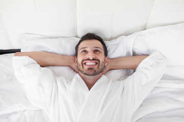 Cheerful young man is waking up after sleeping in the morning. He is yawing and stretching his arms up. His eyes are closed with relaxation. He is lying in the bed