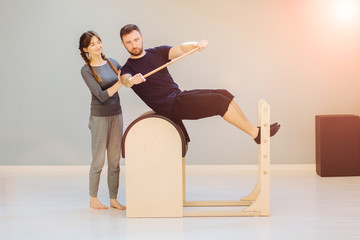 Female instructor of pilates rehabilitation specialist helping to young man patient workout on barrel in studio of rehabilitation center. Healthy life and ecreation after trauma concept.