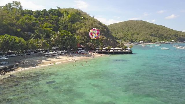 Sea beach aerial landscape. Tourist people swimming in sea and flying on paragliding drone view. Colorful parasail wing pulled by sailing boat in turquoise ocean water.