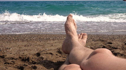 Point of view, first person view. The legs of a man against the background of the sea. A man is lying on the beach, looking at his feet and the sea.