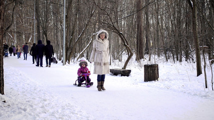 Fototapeta na wymiar Mom pulls a sled with a child. They walk in the winter forest. Daughter in a warm winter overall. A woman of European appearance in a white mink coat.