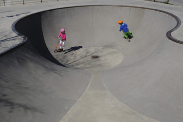 Brother and sister on scooters playing at sunny skate park