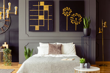 Real photo of a bedroom interior with big, black paintings with golden accents, double bed and plants - obrazy, fototapety, plakaty