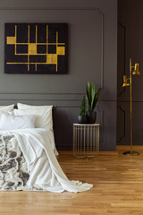 Painting on the grey wall, bedside table with a plant and white bed in a bedroom interior. Real...
