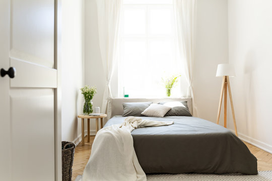 Sunny natural style bedroom interior with a bed in the middle, gray linen and pillows, and a vanilla blanket. A lamp and a stool with fresh flowers next to the bed. Big window in the back. Real photo