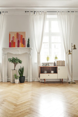 Poster above plants in bright loft interior with gold lamp next to white wooden cupboard. Real photo