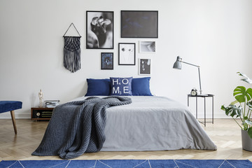 Blanket on bed with blue pillows in white bedroom interior with gallery and lamp on table. Real photo - obrazy, fototapety, plakaty