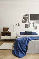 Blue blanket on bed in bright bedroom interior with lamp on cabinet near posters on white wall. Real photo