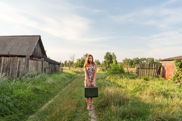 a model girl in a retro dress is holding in her hands a vintage suitcase on an abandoned country...