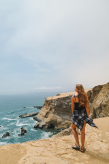 Summer. The girl (blonde) in shorts and top is standing with her back to the sea on a sandy cliff. Peru, Paracas