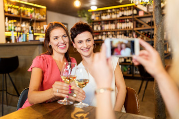 people, technology and lifestyle concept - woman photographing friends drinking wine by smartphone at bar or restaurant