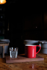 Vertical of red cup of coffee and a glass of iced water on wooden tray on rustic wooden table and wooden chair in background in cafe