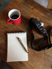 Flat lay of a white notepad and pen red cup of coffee vintage camera on a rustic wooden table in a cafe