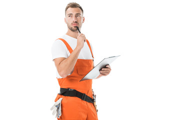 pensive handsome auto mechanic in orange uniform holding clipboard and looking away isolated on white