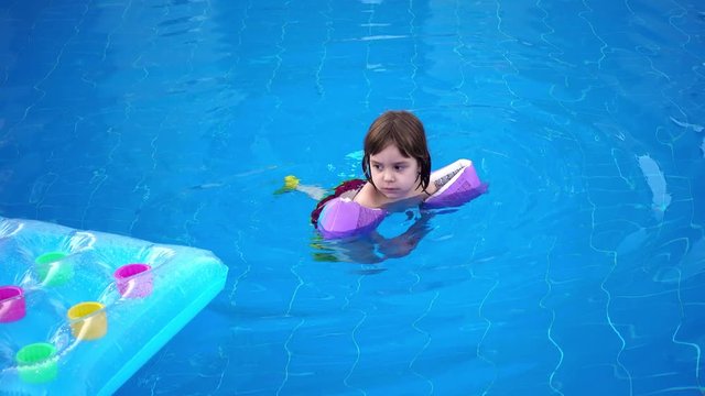 The girl is swimming in the pool. A small child of 4-5 years old, with inflatable armlets, swims in the pool.