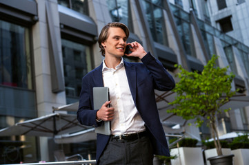 Just signed that contract! Handsome young man carrying his laptop and using his smartphone with smile while standing outdoors