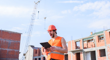Guy in protective helmet stand in front of building made out of red bricks. Man orange vest and helmet works at construction site. Contractor control according to plan. Control construction process