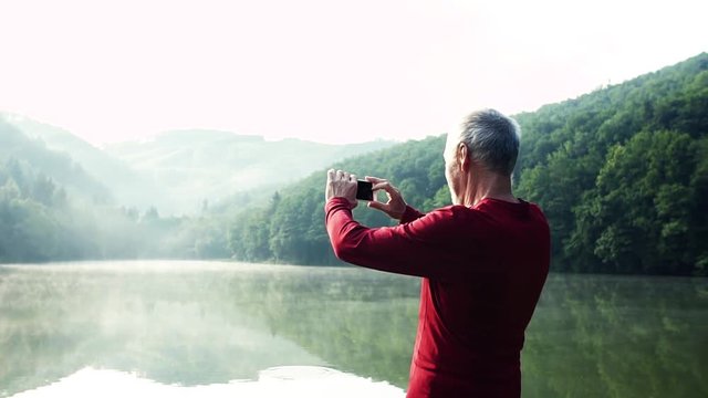 Senior man standing by the lake outdoor in foggy morning in nature, taking photograph.