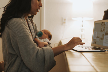 Mother using a computer and holding her baby - obrazy, fototapety, plakaty
