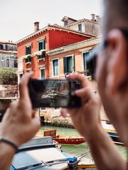 A man holding a mobile phone and taking a photo on the phone screen shows the channel and the boat in Venice
