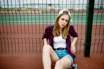 A pretty blond girl wearing checkered shirt, white cap and denim shorts is sitting leaned on the guardlattice on the sports field. Sport and cool style.