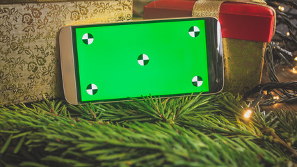 CLoseup toned image of mobile phone with empty green screen for inserting your text or picture against Christmas decorations and gifts