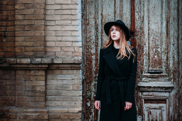 Blonde beautiful girl with long hair, in black coat in hat, stands on the background of vintage antique old wooden door old brick house. For concept design.