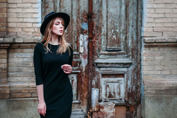 Blonde beautiful girl with long hair, in black dress in hat, stands on the background of vintage antique old wooden door old brick house. For concept design.