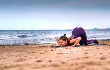 brown-haired woman performs a yoga complex of suray namaskara on the seashore in the sand on a clear day