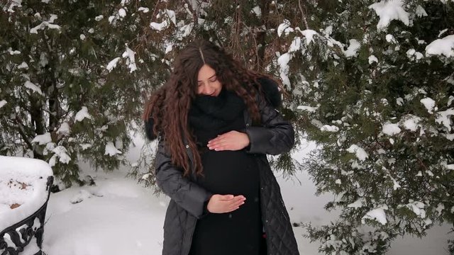 A beautiful pregnant woman is stroking her big belly in a winter park among lots of snow. Pregnancy concept. Portrait of expectant mother.