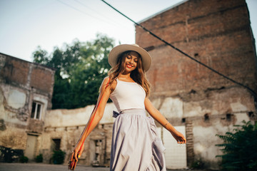 Young woman in hat goes at city street next to brick wall of building.