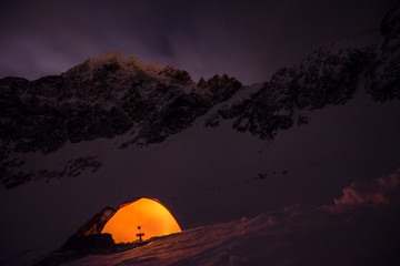 Orange tent under starry sky and milky way. Extreme and adventure winter camping under the stars in winter alpine like landscape. Epic bivi spot. Mountaineering and alpinism tent shining in the night.