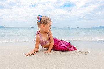 Little beautiful girl dressed in swimsuit as a mermaid sits on the seashore