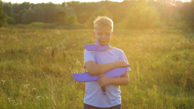 Closeup sunny portrait of handsome young kid holding and enjoying his new toy blue plane. Boy playing outdoors in sunset meadow on warm summer evening. Real time full hd video footage.