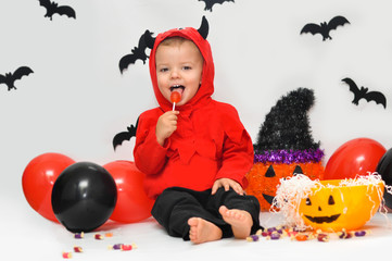 Laughing cute baby boy in costume devil sitting on a background of scenery for Halloween and eating red lollipop.