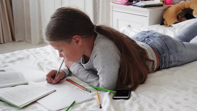 Little school girl lying on bed and doing homework