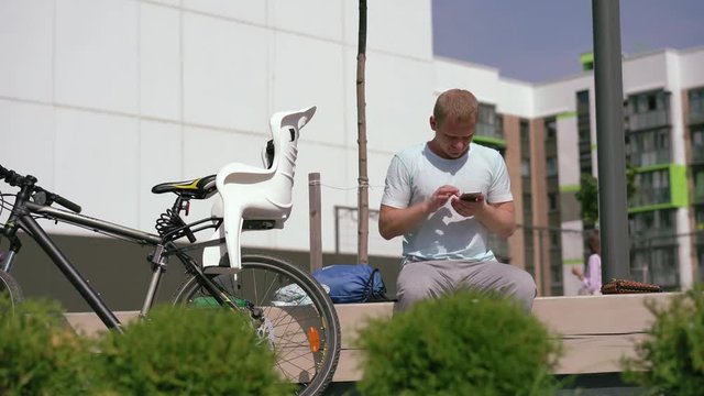 modern life in a big world - young man sits in the city on a bench and rewrites in smartphone