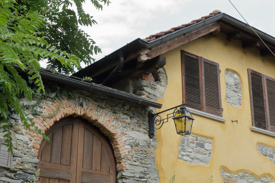 A yellow countryside stone house with two windows with louvres, a wooden front door next to a tree and a street lantern
