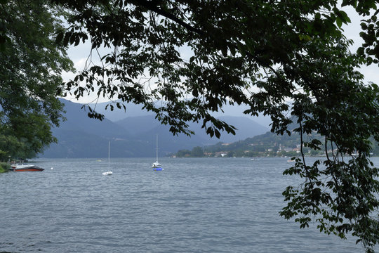 A landscape of Lake Orta, in northern Italy, in a leaves and branches frame, with two sailing boats and mountains on the background