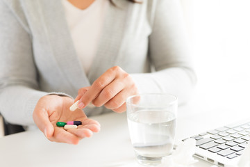 Young business woman sick in the office, hand holding pills and water glass. pills, tissue and laptop on table. Healthcare And Medical concept.