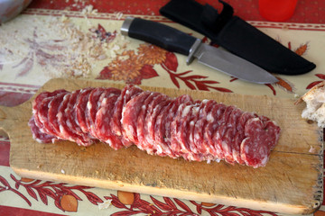 Slices of red Italian salami on a wooden cutting board on a table with red and white tablecloth and a knife with is sheath on a side