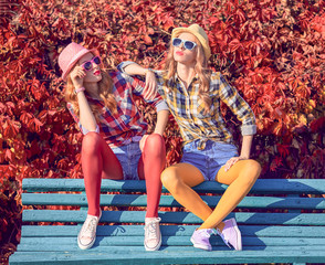 Autumn Fashion. Two Young Hipster Woman Relax sitting on bench in Fall Park. Beautiful Girl Friends Enjoy Nature wearing plaid shirt. Urban Outdoor. Model in Stylish Trendy Outfit